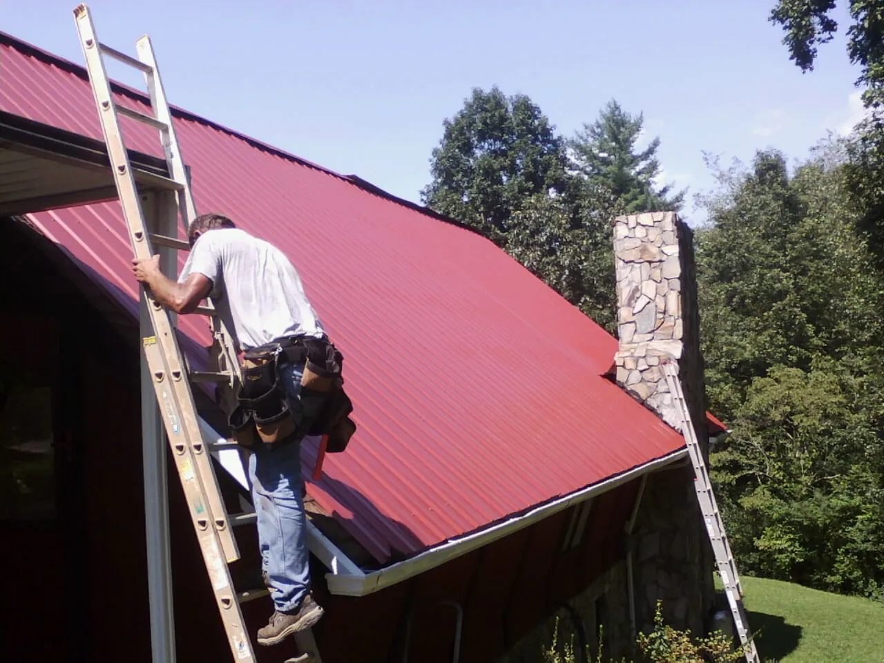 Roofer on a ladder beside a red metal roof and stone chimney — Soffit & Fascia Repair by Roofing Brothers in Puyallup